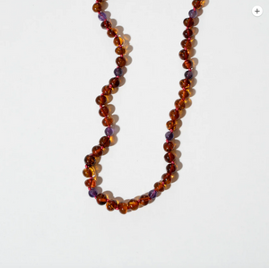 Bernsteinkette „Amethyst“ von Nirrimis aus poliertem Bernstein und Amethyst, handgeknüpft mit Silberverschluss, auf Leinenbeutel präsentiert.